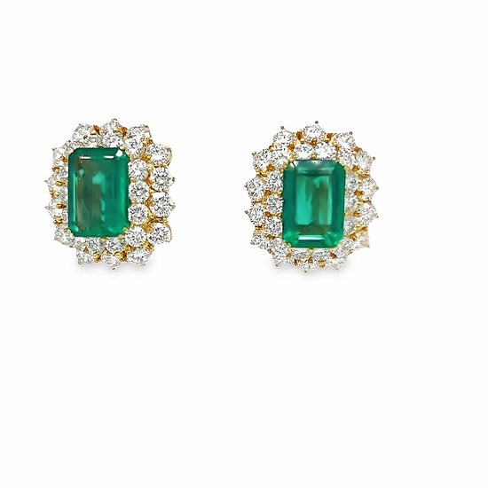 Close up of Emerald and Diamond Stud Earrings. Set in 18k yellow gold, these studs feature emerald cut green emerald gemstones and a starburst halo of round white diamonds.